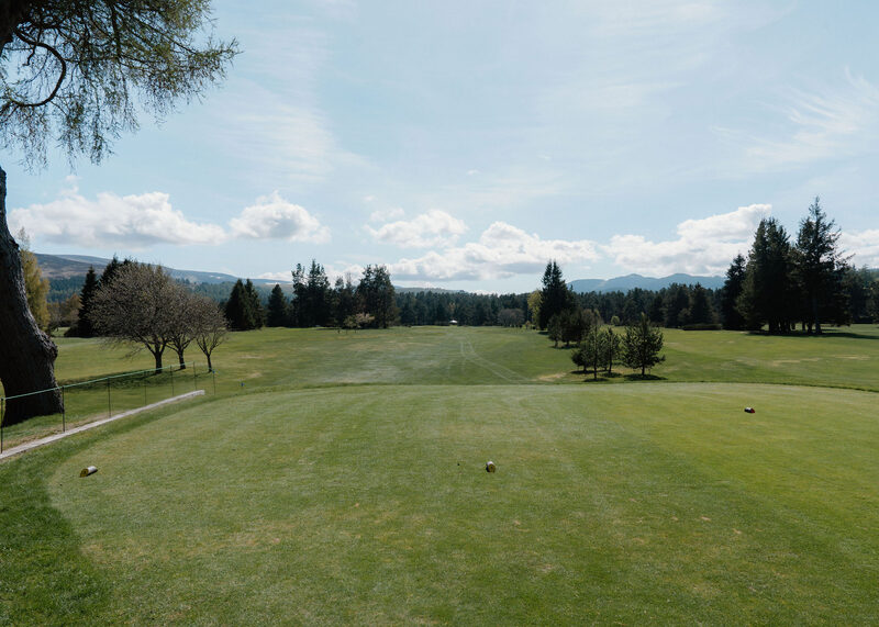 A tee looking down a long straight fairway between trees towards distant hills.