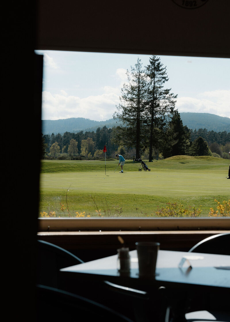 A golfer putting on a green with a red flag, seen through the clubhouse window.
