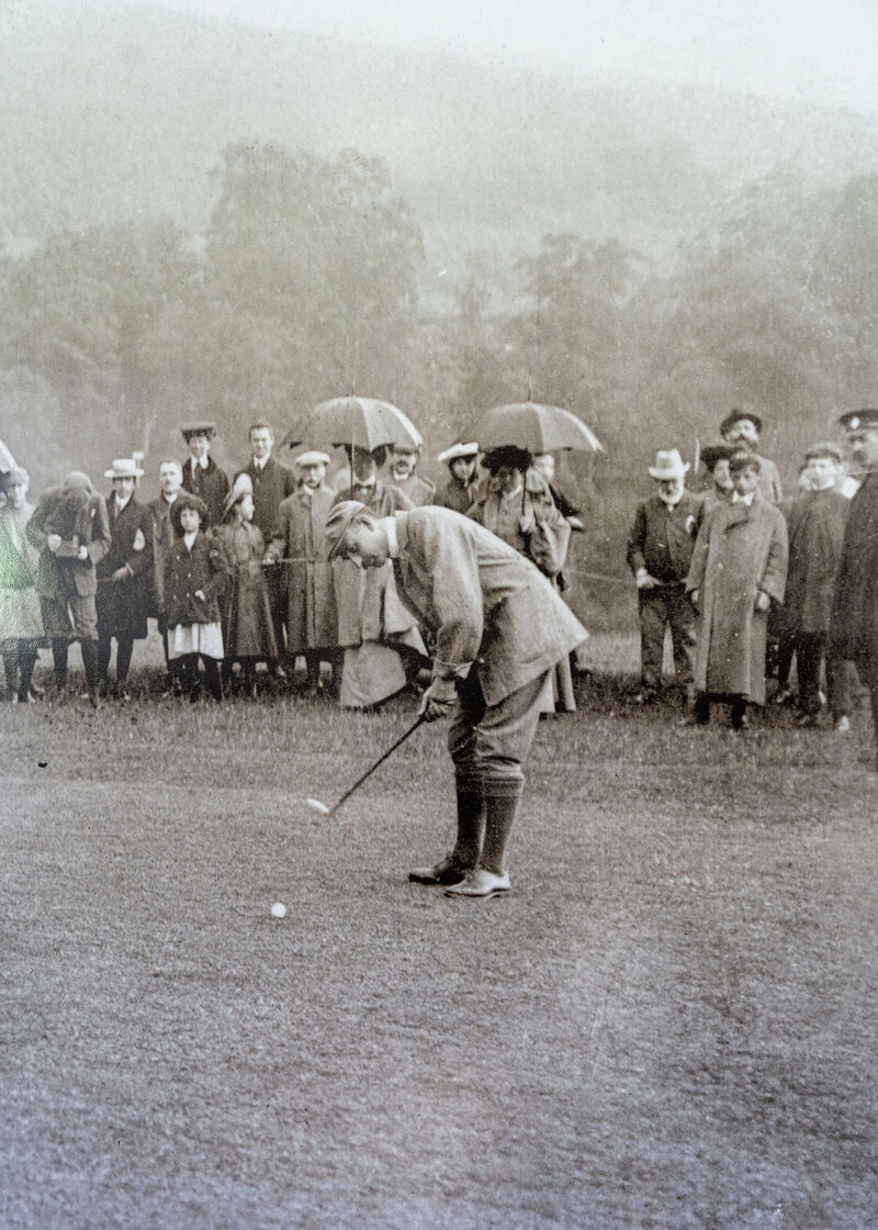 An old photograph of a golfer in plus fours putting in the rain, watched by a crowd under umbrellas.