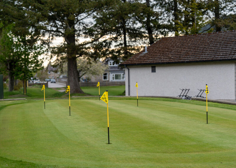 A putting green with yellow flags beside a white harled building, with trolleys and houses beyond.