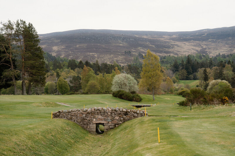 A stone bridge over a grassy ditch on the course, with yellow posts and heather hills behind.