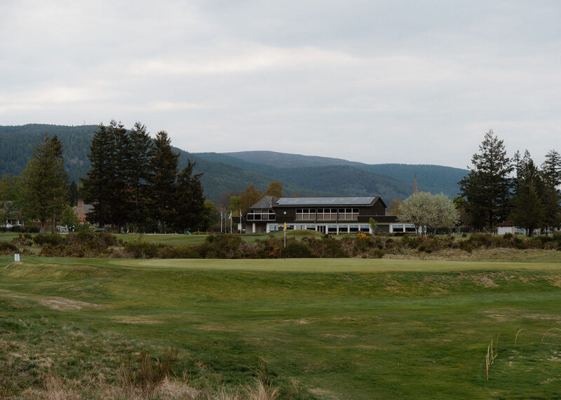 The clubhouse of Ballater Golf Club across a green, with flagpoles, blossom and forested hills.