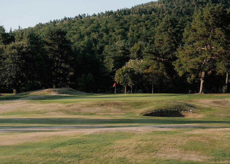 A green with a red flag among grassy mounds and a bunker, below a pine-covered hill.
