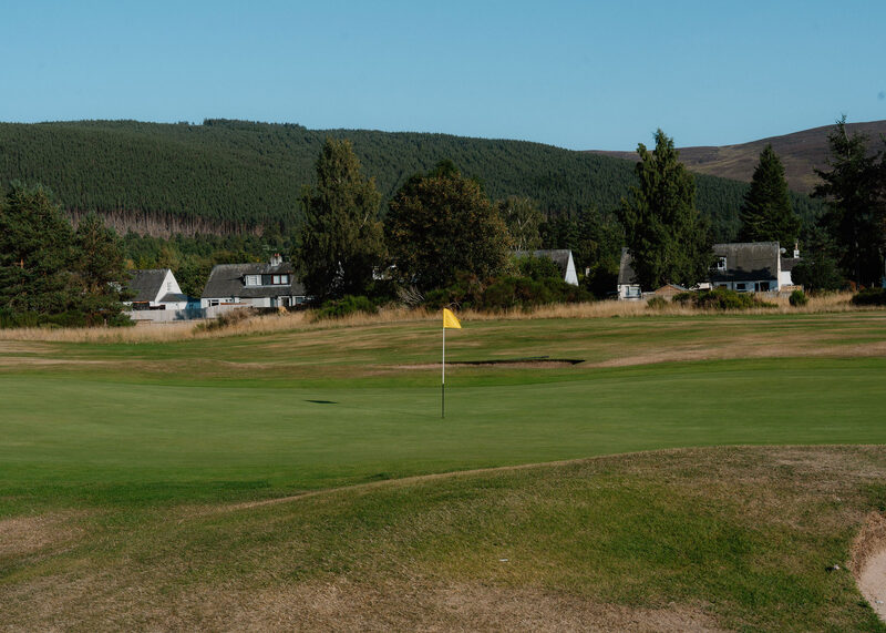 A green with a yellow flag, with white cottages and a forested hill beyond.