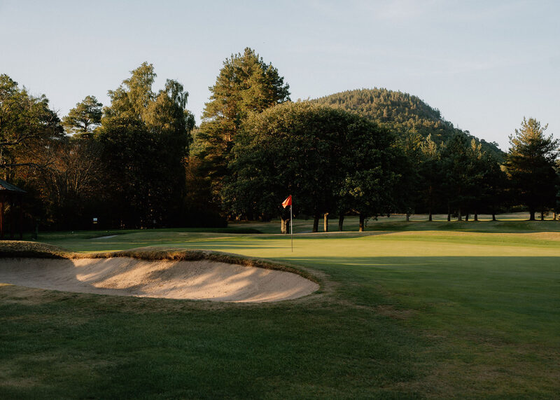 A green with a red flag beyond a bunker, with a rounded wooded hill behind.