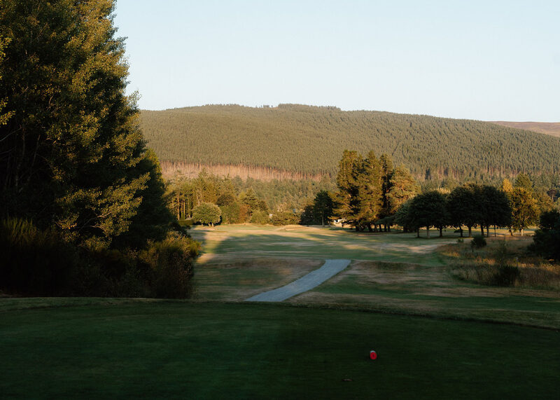 A tee with a red marker above a path down to the fairway, below a forested hill in evening light.