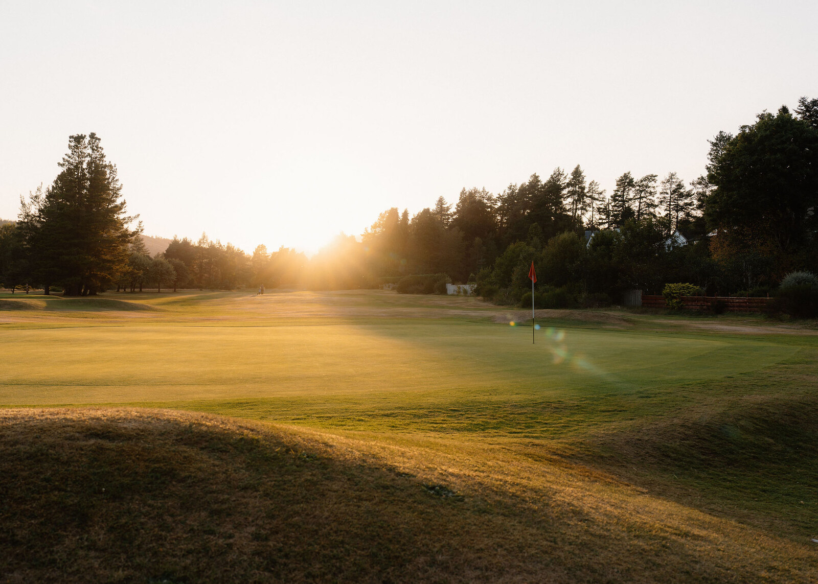 Low sun over a green with a red flag, beyond grassy mounds.