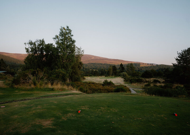 A tee with red markers above a heather gully at dusk, with a heather hill behind.