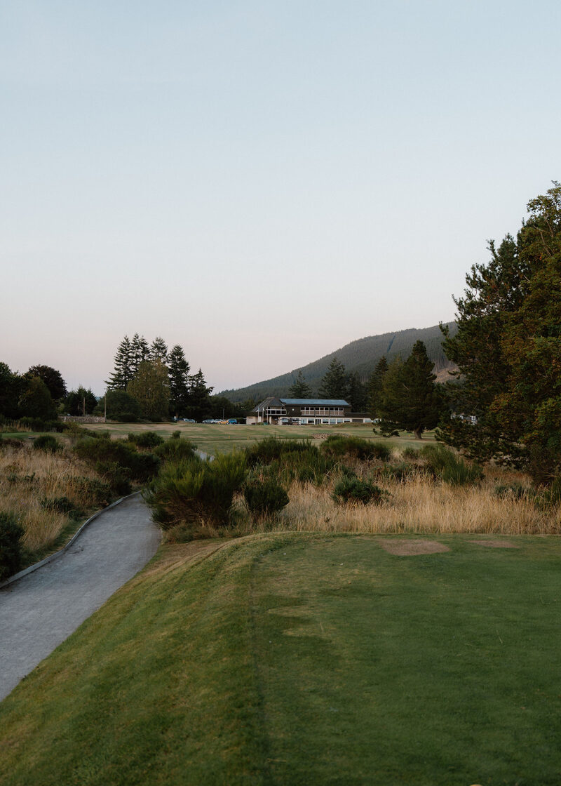 A path leading across the course to the lit clubhouse at dusk, below a hill.