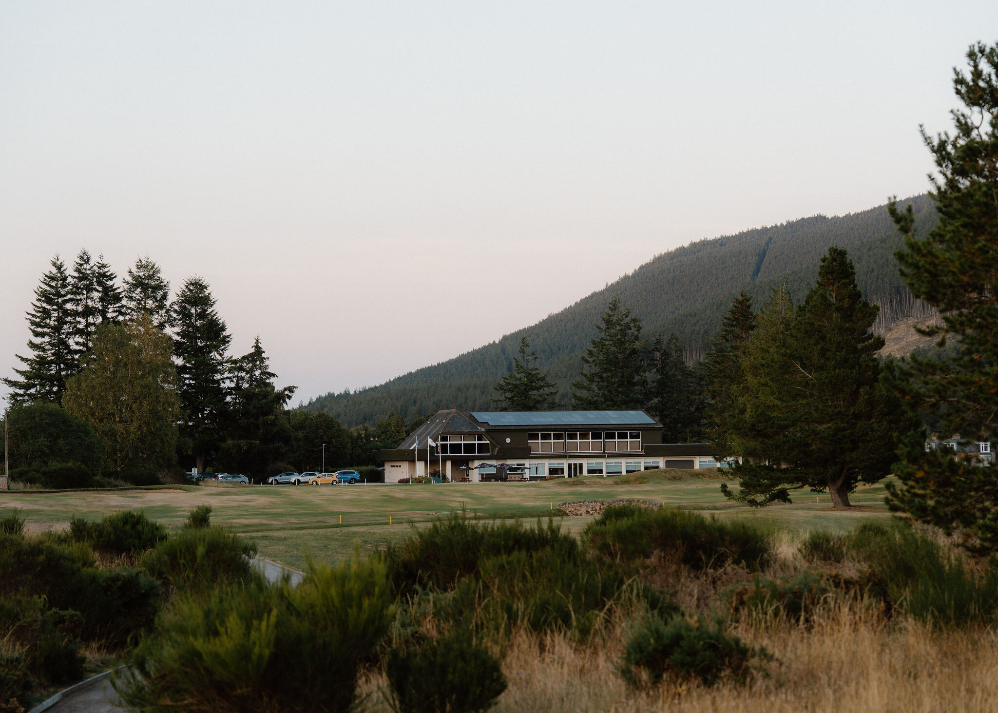 The clubhouse of Ballater Golf Club across the course at dusk, beyond gorse and broom, with a forested hill behind.