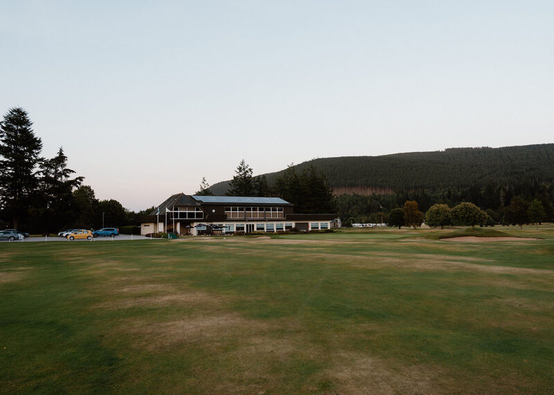 The clubhouse and car park across a fairway at dusk, with a forested hill behind.