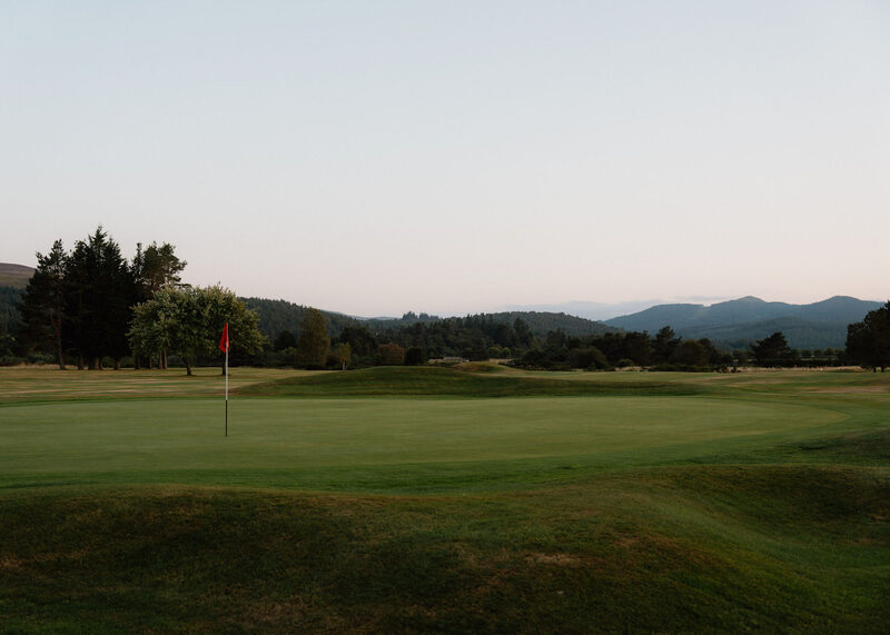 A raised green with a red flag at dusk, with hills on the horizon.