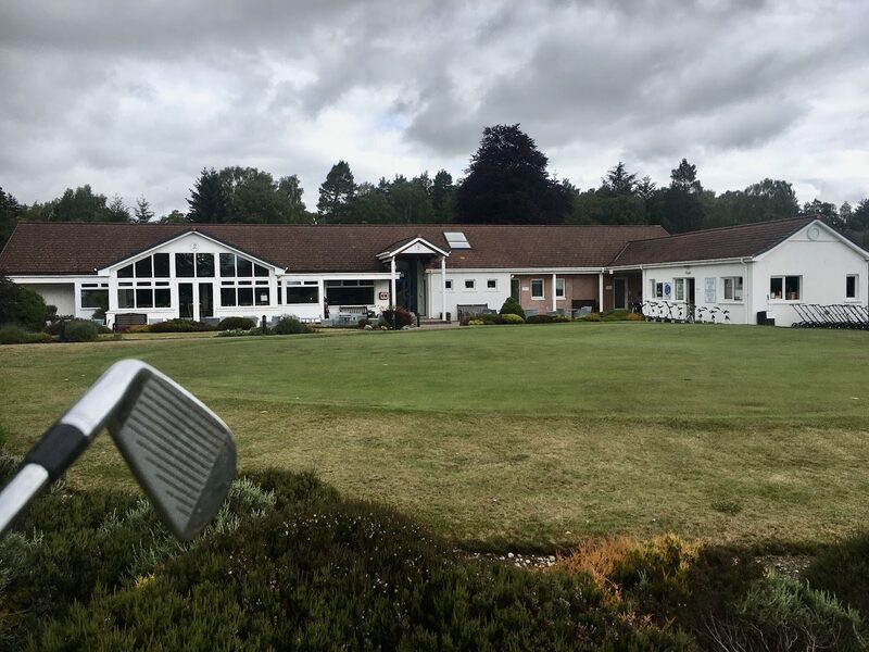 The white clubhouse with its tall gabled window, seen across a green, with trolleys lined up outside the right-hand wing and an iron held up in the foreground.