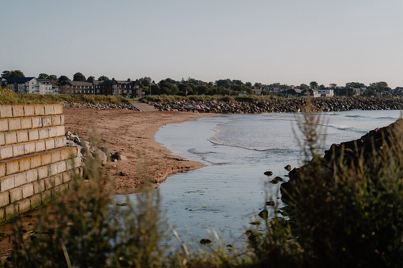 A curving sandy beach beneath rock armour, houses lining the shore in the distance.