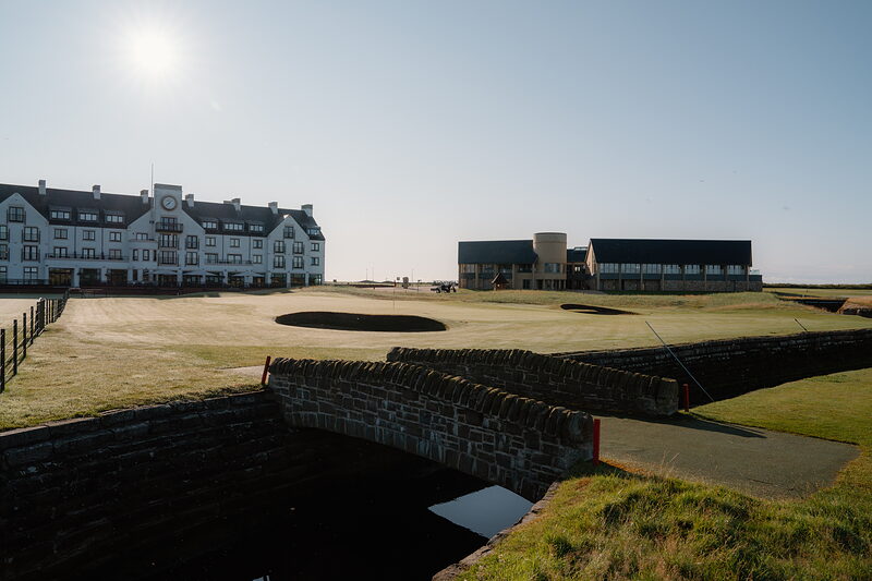 A stone footbridge crossing a burn before a row of hotel and clubhouse buildings lit by low sun.