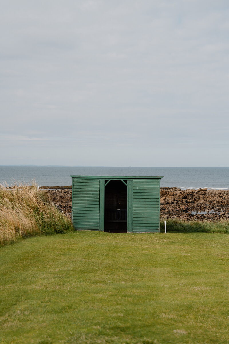 A green wooden beach hut standing on grass beside a rocky shoreline and the sea