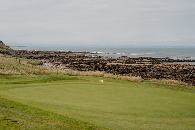 A putting green bordered by rocky coastline and the sea, a yellow flag marking the hole