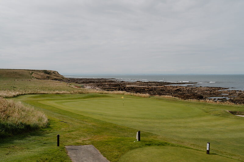 A putting green bordered by rocky coastline and the sea, a red flag marking the hole