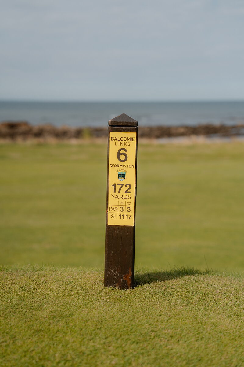 A wooden marker post for Balcomie Links hole 6, Wormiston, giving the yardage and par beside the sea
