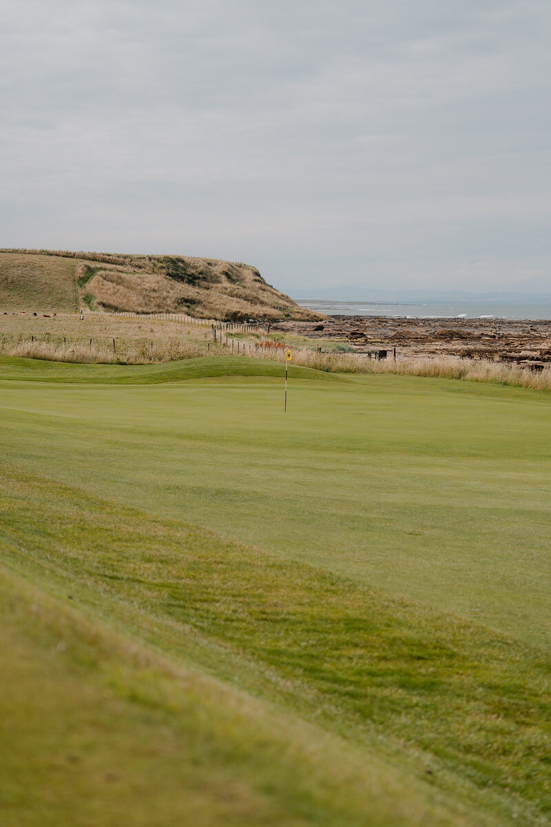A green with a yellow flag bordered by a wooden fence, rocky shoreline and the sea beyond