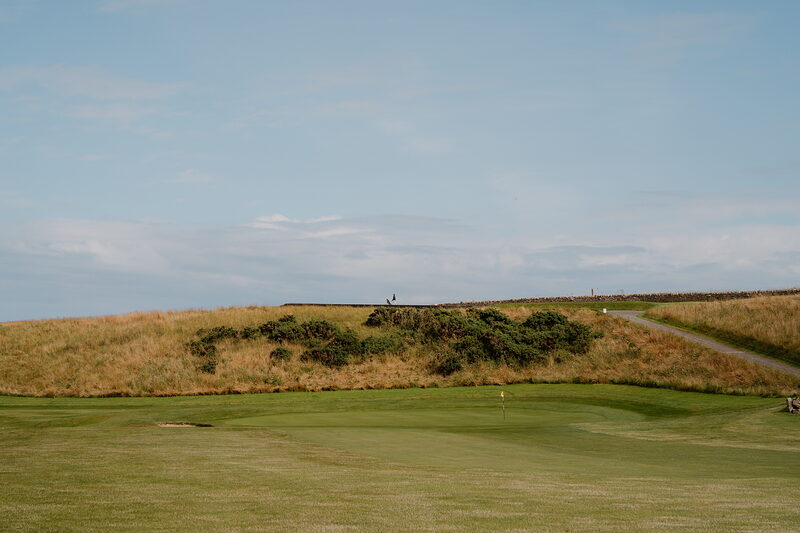 A gravel path winding past dune bushes towards a green, figures visible in the distance