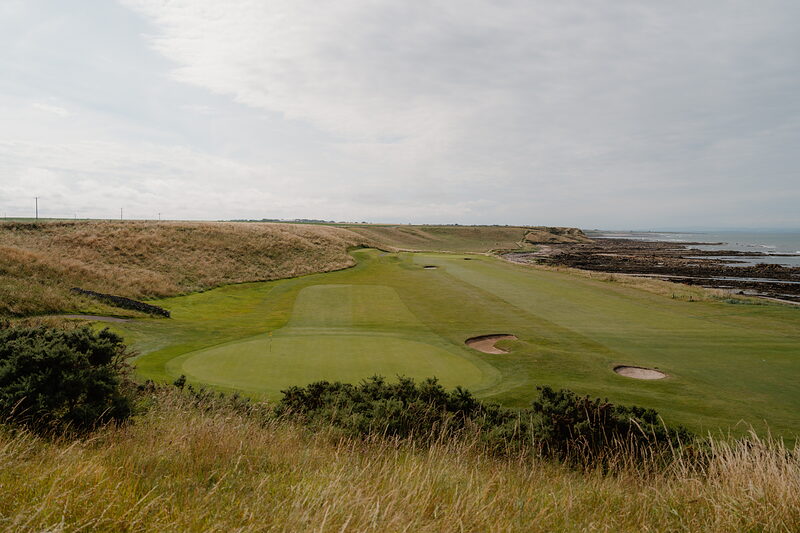A green bordered by a bunker overlooking rocky coastline and the sea