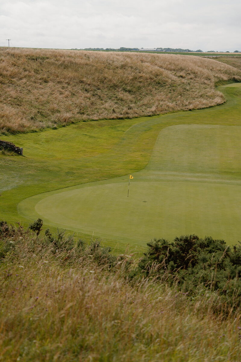 A green with a yellow flag nestled between grassy hillocks, gorse bordering the fairway