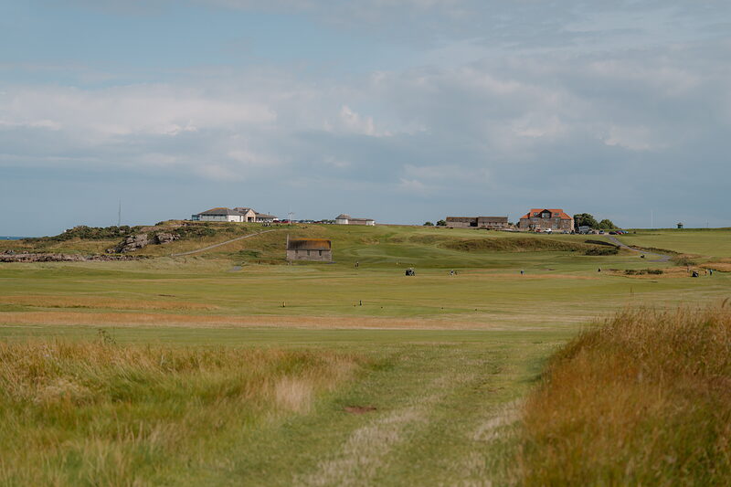 A links course running towards farm buildings on a headland, a worn path leading through the rough