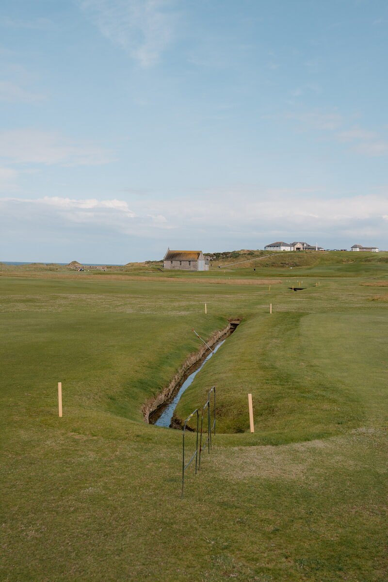 A stream winding through a fairway bunker before thatched buildings on a distant rise