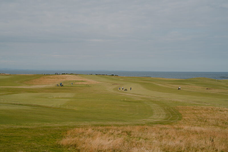 Golfers walking a fairway towards distant dunes, the sea visible on the horizon