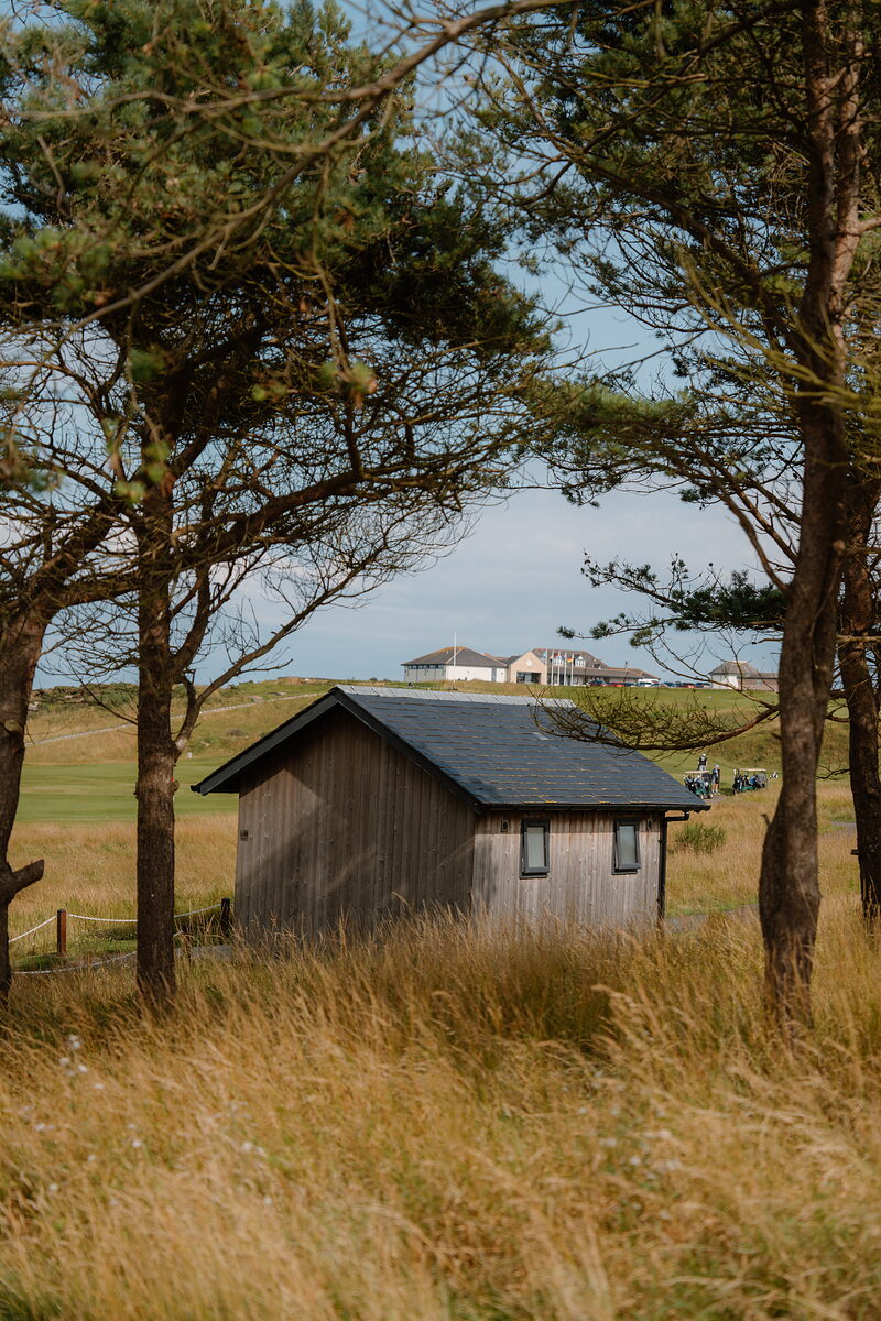 A dark timber shelter set among wind-bent pines, a clubhouse visible on the rise behind