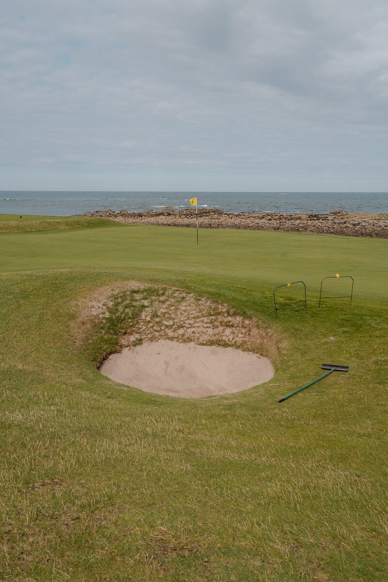 A bunker beside a green marked by a yellow flag, the sea and rocky shoreline visible beyond