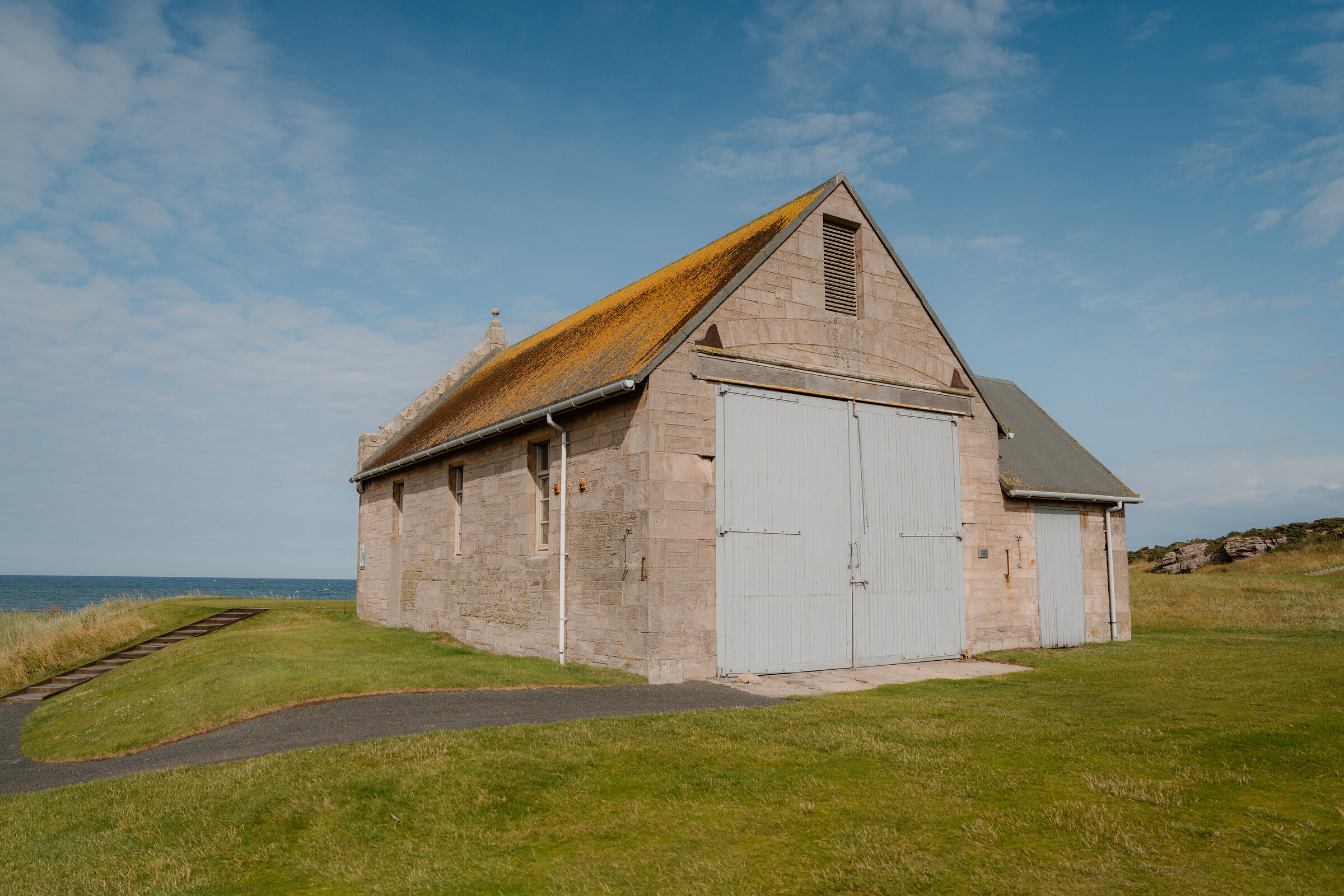 A sandstone boathouse with white doors standing on a grassy bank beside the sea