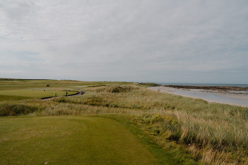 Distant figures crossing a wooden bridge beside a sandy shoreline, dune grass framing the fairway