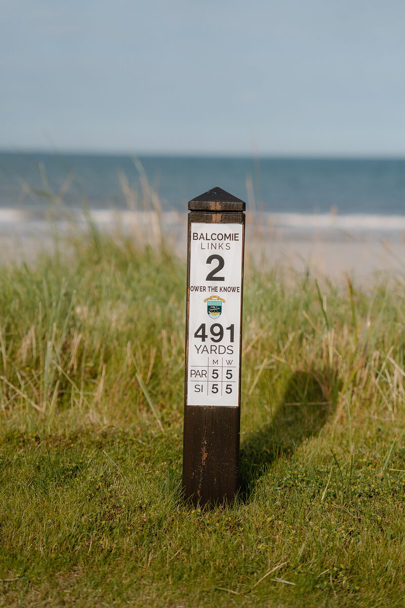 A course marker post reading Balcomie Links, hole 2, 491 yards, standing above a sandy beach