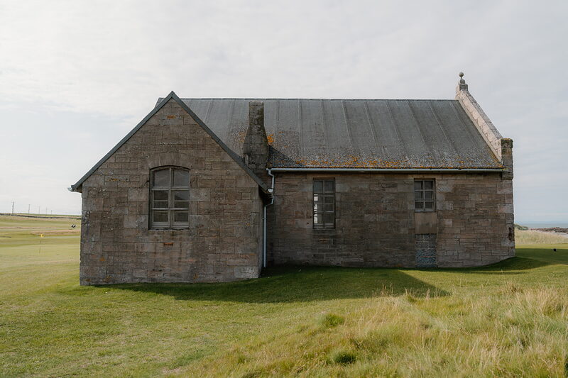 A weathered sandstone building with a corrugated roof standing alone on a fairway of long grass