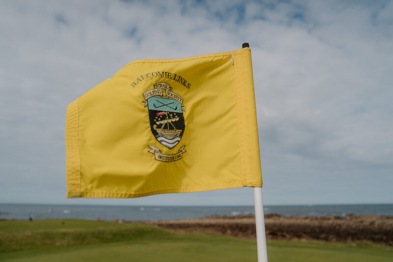 A yellow flag bearing the Balcomie Links crest fluttering above a green, the sea stretching behind