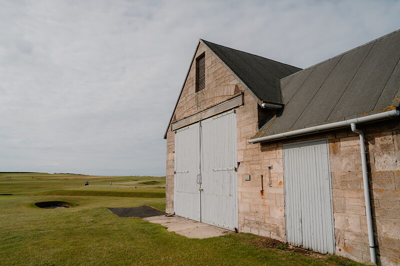 A sandstone building with white sliding doors and a slated roof standing on a fairway of long grass