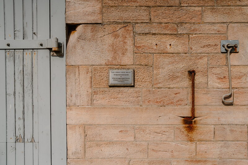 A weathered brass plaque mounted on a sandstone wall beside a timber door and drainpipe