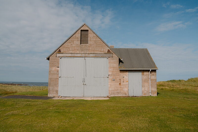 A sandstone boathouse with white sliding doors and a slated roof standing on a grassy bank beside the sea