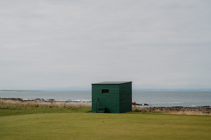 A green timber shelter standing alone on a fairway overlooking the sea
