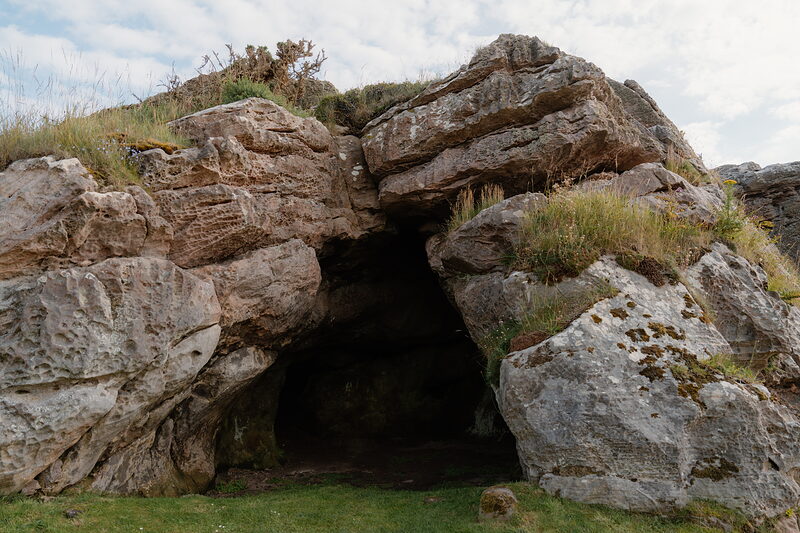 A rocky cave entrance framed by grass and gorse growing along its rim