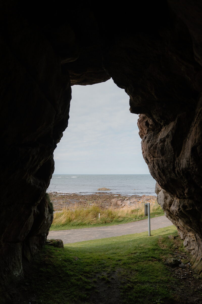 A sea view framed by the mouth of a rocky cave, a path and marker post visible on the shore beyond
