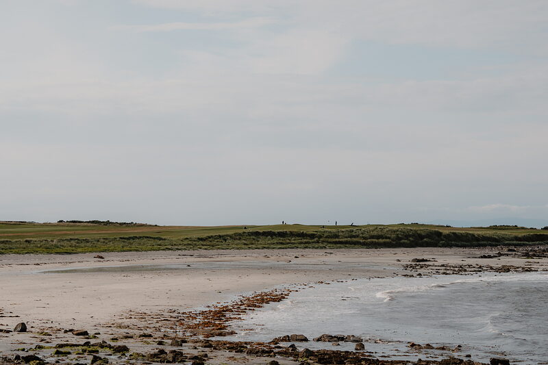 A curving sandy beach beneath a grassy headland, distant figures walking along the shore