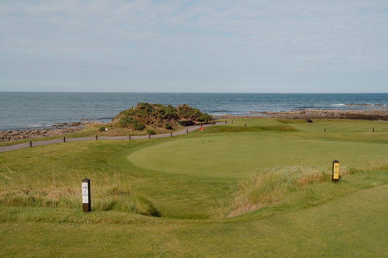 A green beside the sea, a rocky headland covered in gorse rising behind it