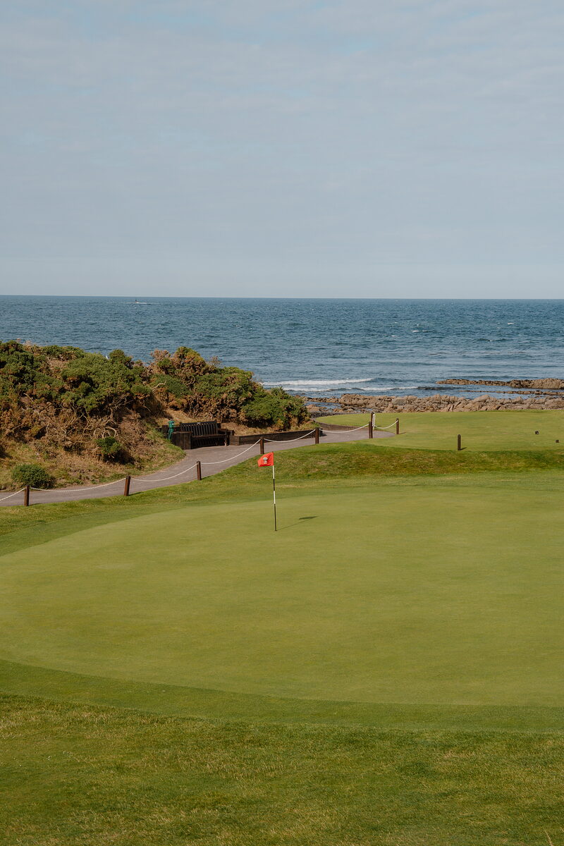 A green marked by a red flag overlooking the sea, a path curving along the rocky shore