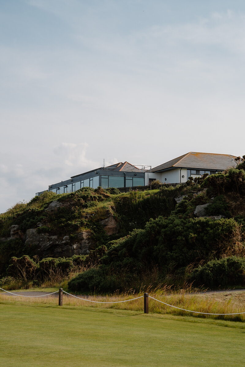 A clubhouse perched on a rocky headland covered in gorse, a rope fence lining the fairway below