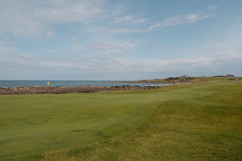 A green marked by a yellow flag beside a rocky shoreline, the sea stretching to the horizon