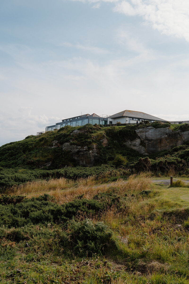 A clubhouse perched on a rocky cliff edge above gorse-covered dunes