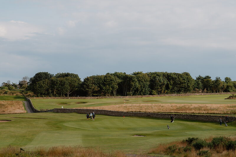Golfers putting on a green bordered by a stone wall and woodland